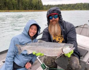 A bearded fishing guide smiles while holding a salmon for a guest during an all-inclusive Alaska fishing trip.