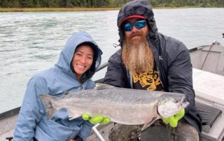 A bearded fishing guide smiles while holding a salmon for a guest during an all-inclusive Alaska fishing trip.